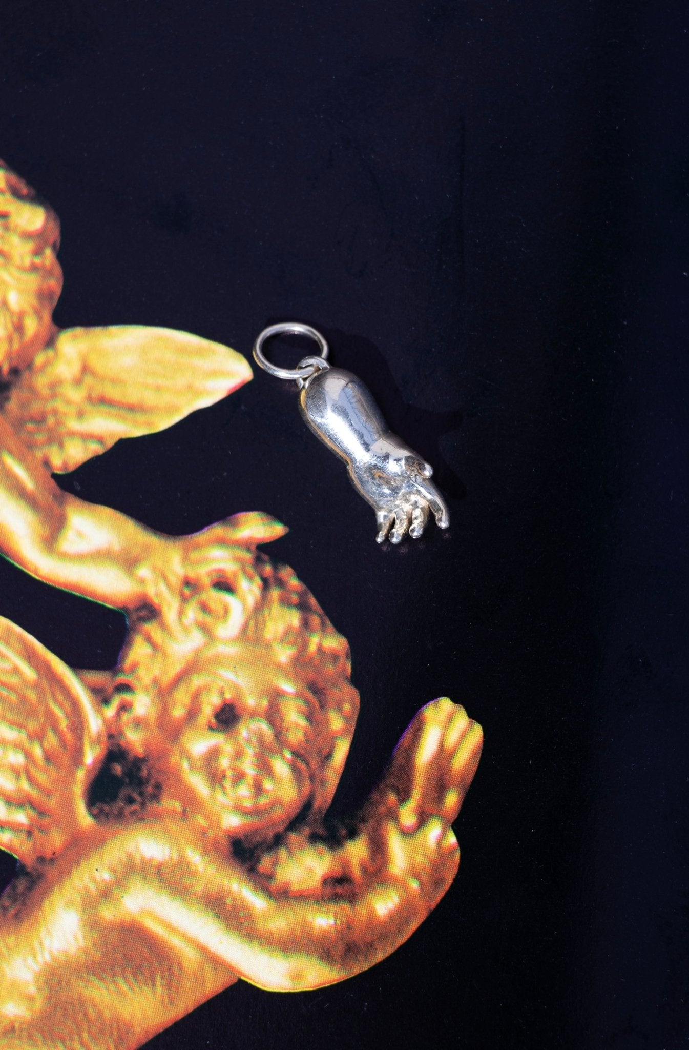 large sterling silver baby hand charm on a black background with a gold cherub catching it - queer and trans fine jewelry by lettuce goods / 14karatdoll
